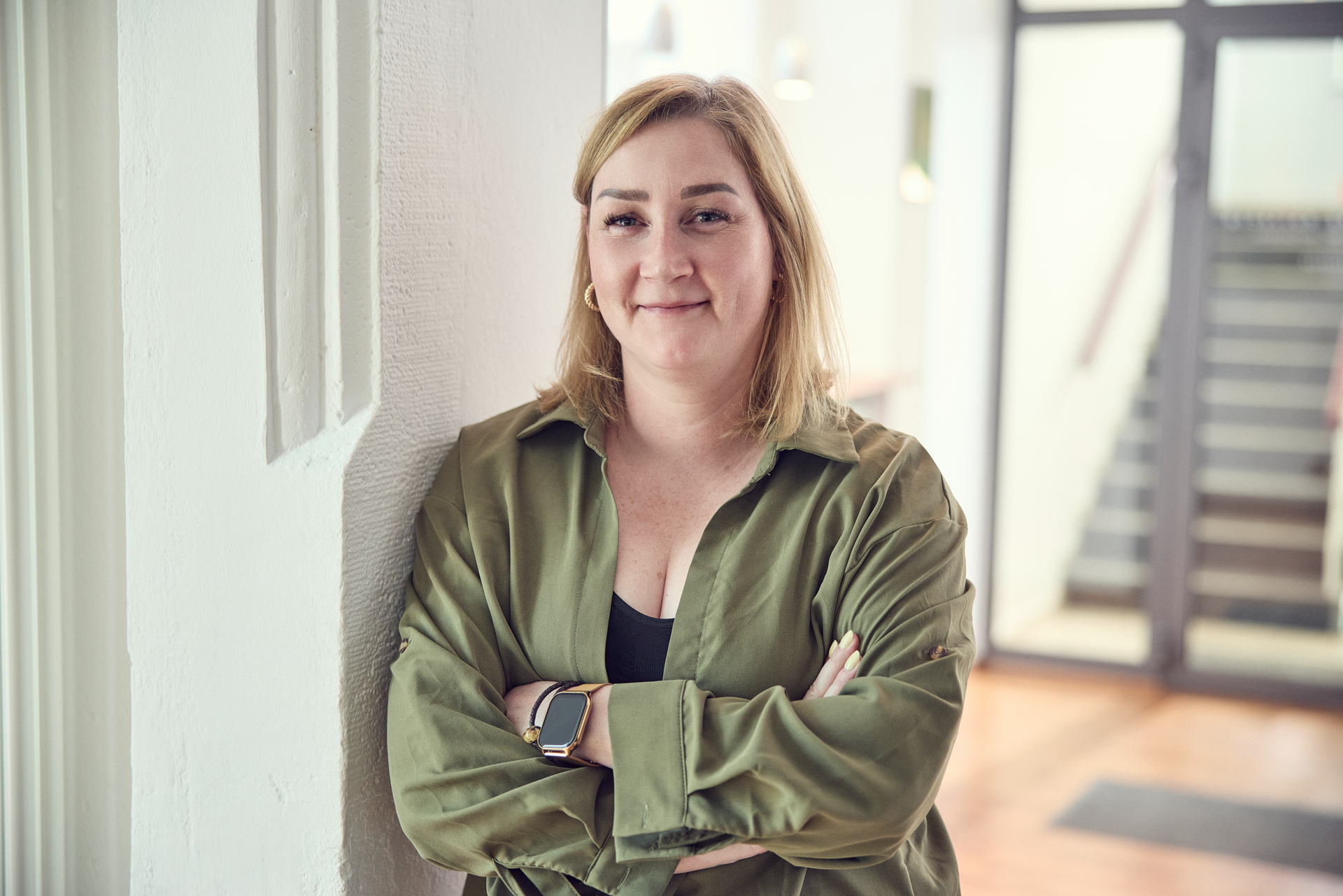 Eine Person mit blonden Haaren steht an einer Wand und lehnt mit verschränkten Armen. Sie trägt eine grüne Bluse und hat einen selbstbewussten Gesichtsausdruck. Im Hintergrund sind unscharfe Elemente eines modernen Innenraums erkennbar.