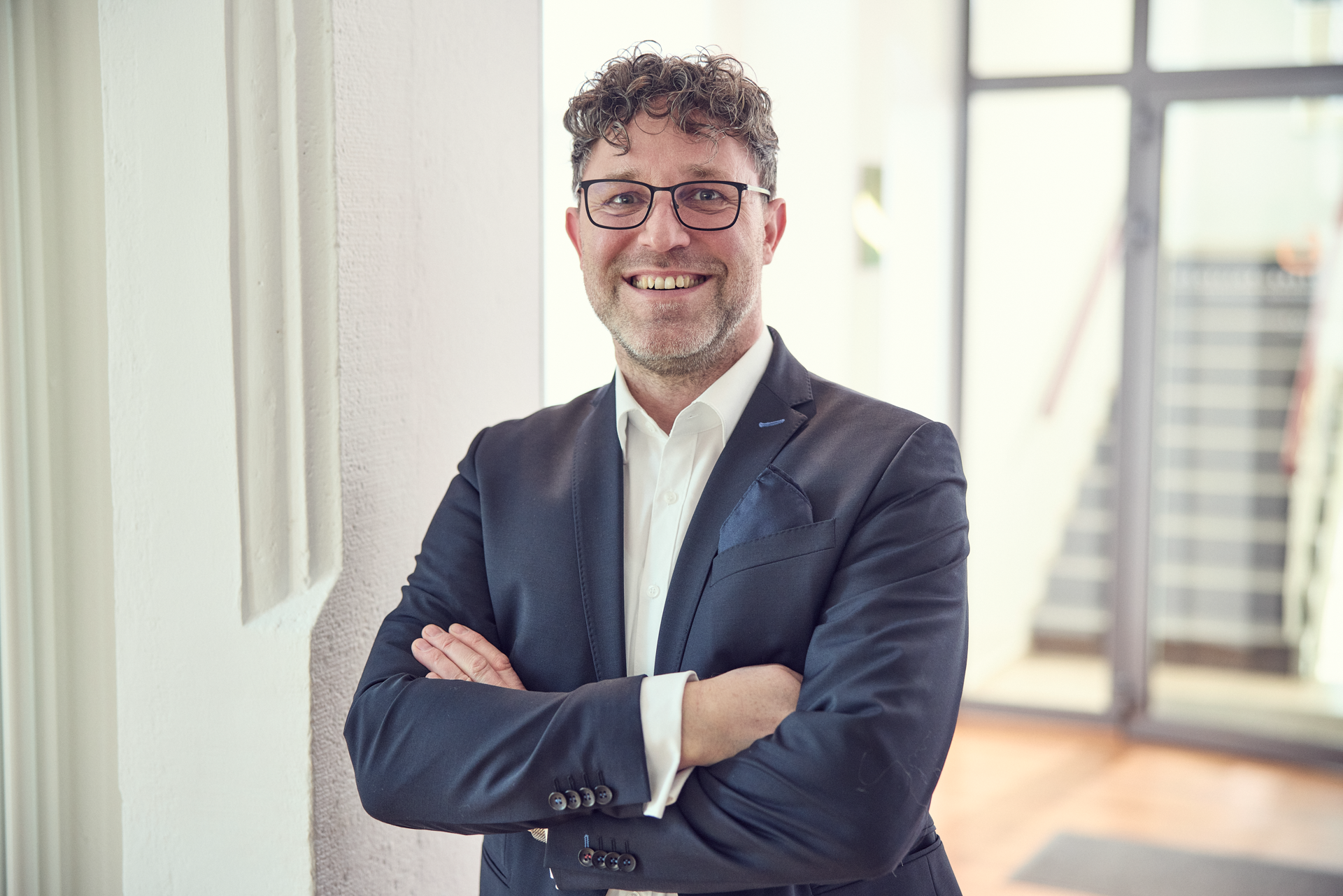 Ein Mann mit lockigem, dunkelbraunem Haar und Brille steht mit angewinkelten Armen und lächelt freundlich. Er trägt einen schwarzen Anzug und ein weißes Hemd. Im Hintergrund sind Treppen und ein heller Raum zu sehen.