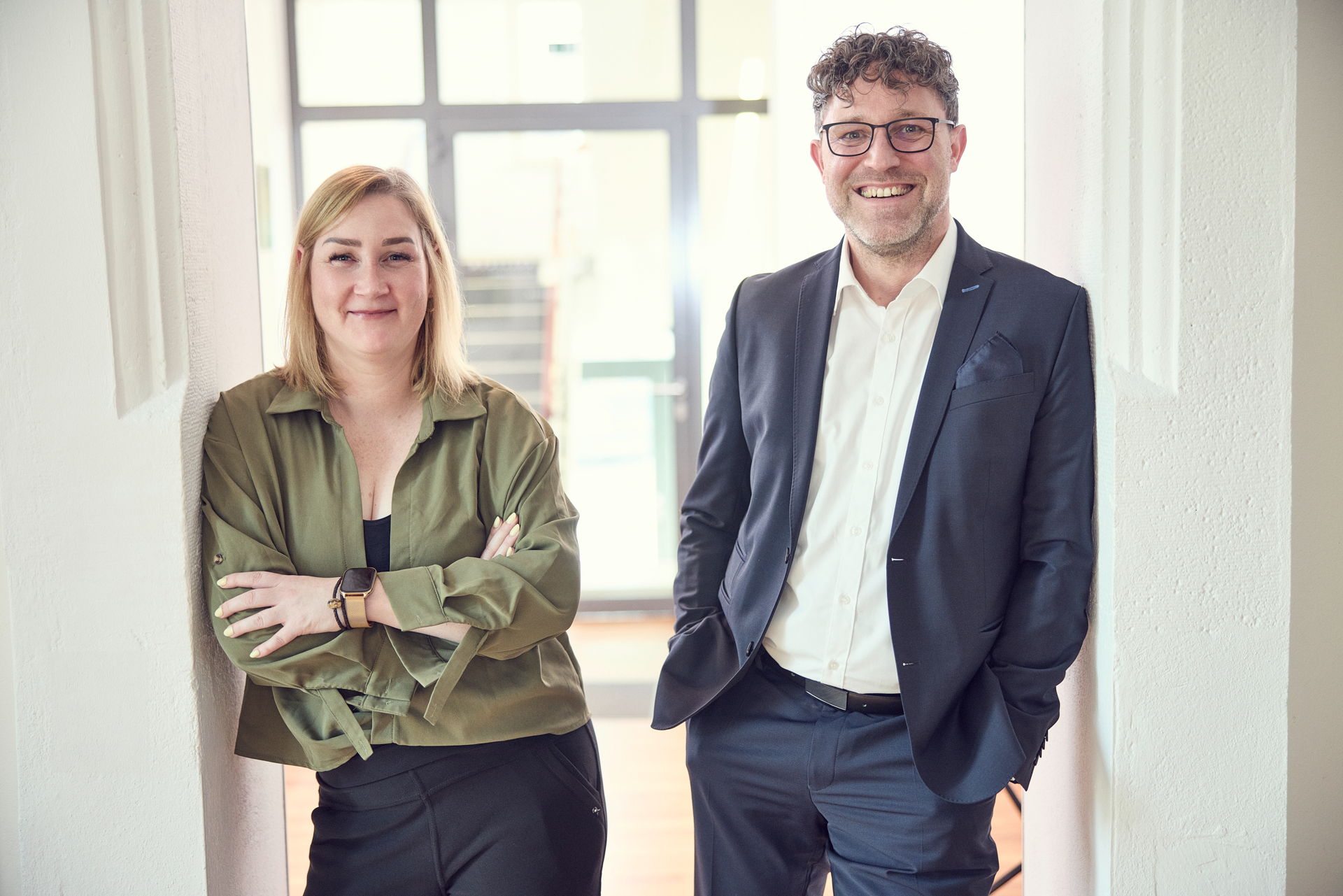 Zwei Personen stehen in einem hellen, modernen Raum; die Frau trägt eine grüne Bluse und der Mann einen Anzug, beide lächeln freundlich in die Kamera.