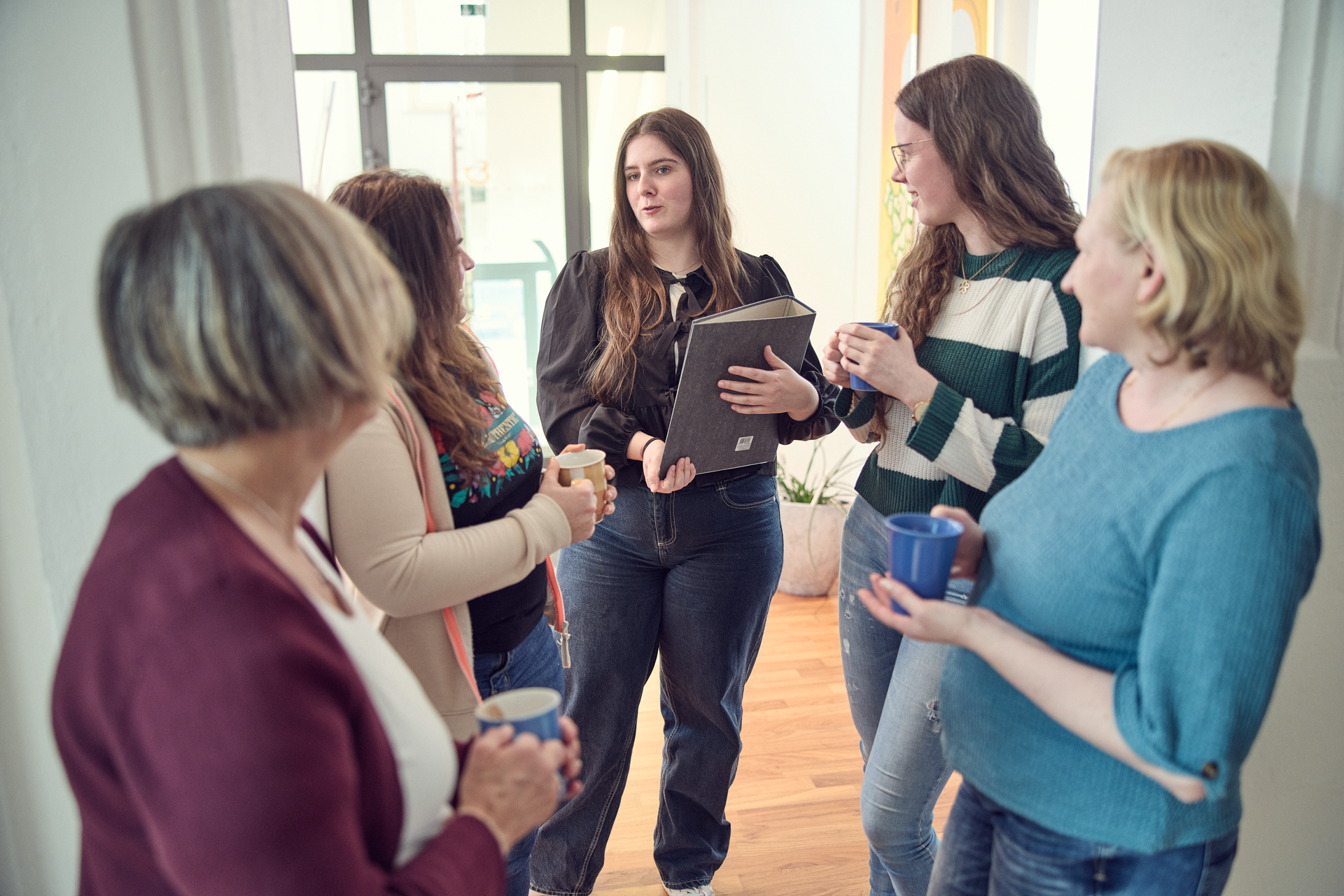 Sechs Frauen stehen in einem hellen Raum zusammen, während eine von ihnen ein Notizbuch hält und spricht. Die anderen halten Tassen in den Händen und hören aufmerksam zu. Pflanzen sind im Hintergrund sichtbar.