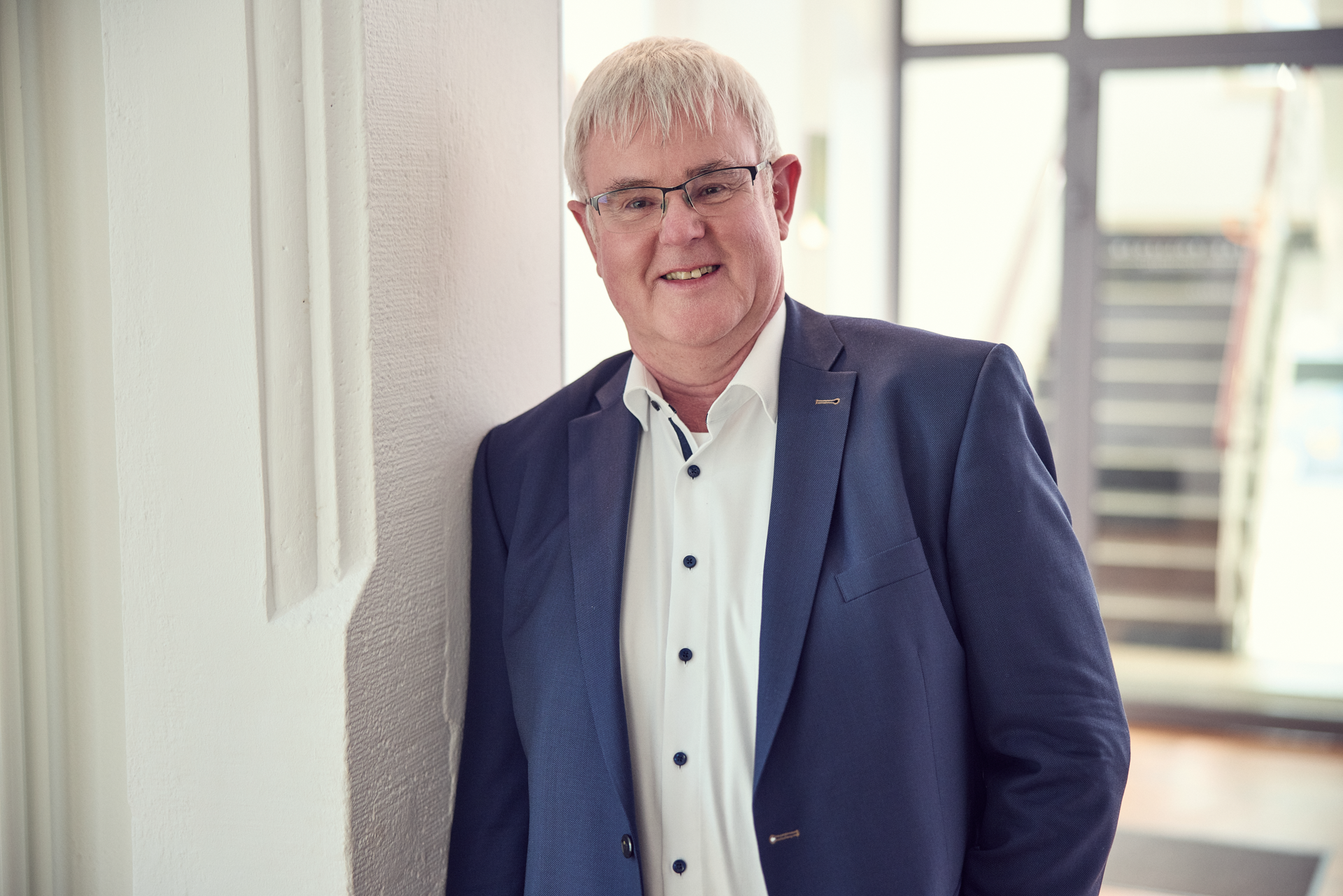 Ein Mann mit kurzen, grauen Haaren und Brille lehnt lächelnd an einer Wand und trägt einen blauen Anzug mit einem weißen Hemd. Im Hintergrund sind Treppen und ein heller, moderner Raum sichtbar.