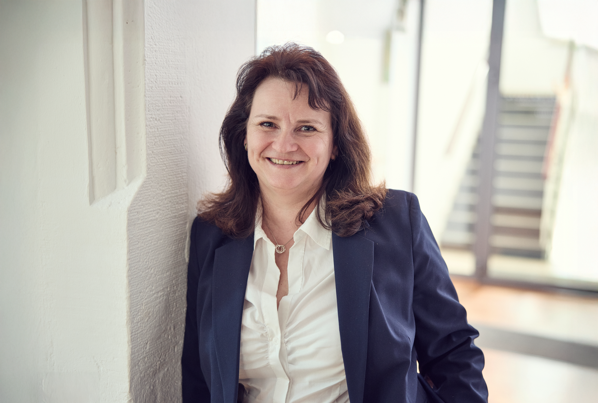 Eine lächelnde Frau mit langen, dunklen Haaren steht an einer Wand und trägt ein schwarzes Blazer und ein weißes Hemd. Im Hintergrund sind Treppen und ein heller Flur zu sehen.