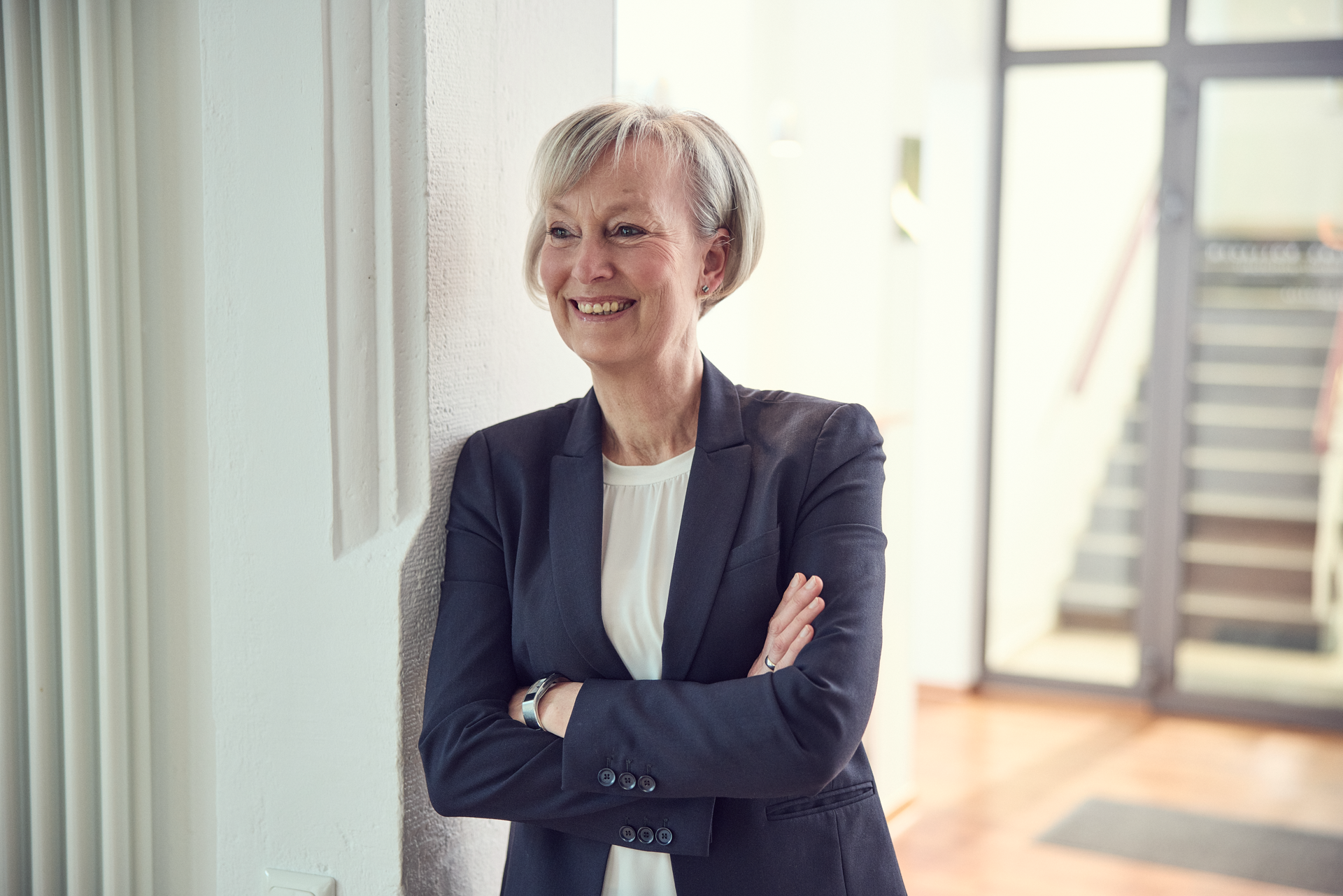 Eine lächelnde Frau mit kurzen, blonden Haaren steht an einer Wand, die Arme mühelos verschränkt, während sie in einem modernen Innenraum mit Holzfußboden und Treppe im Hintergrund positioniert ist. Sie trägt einen schwarzen Blazer über einem weißen Oberteil.