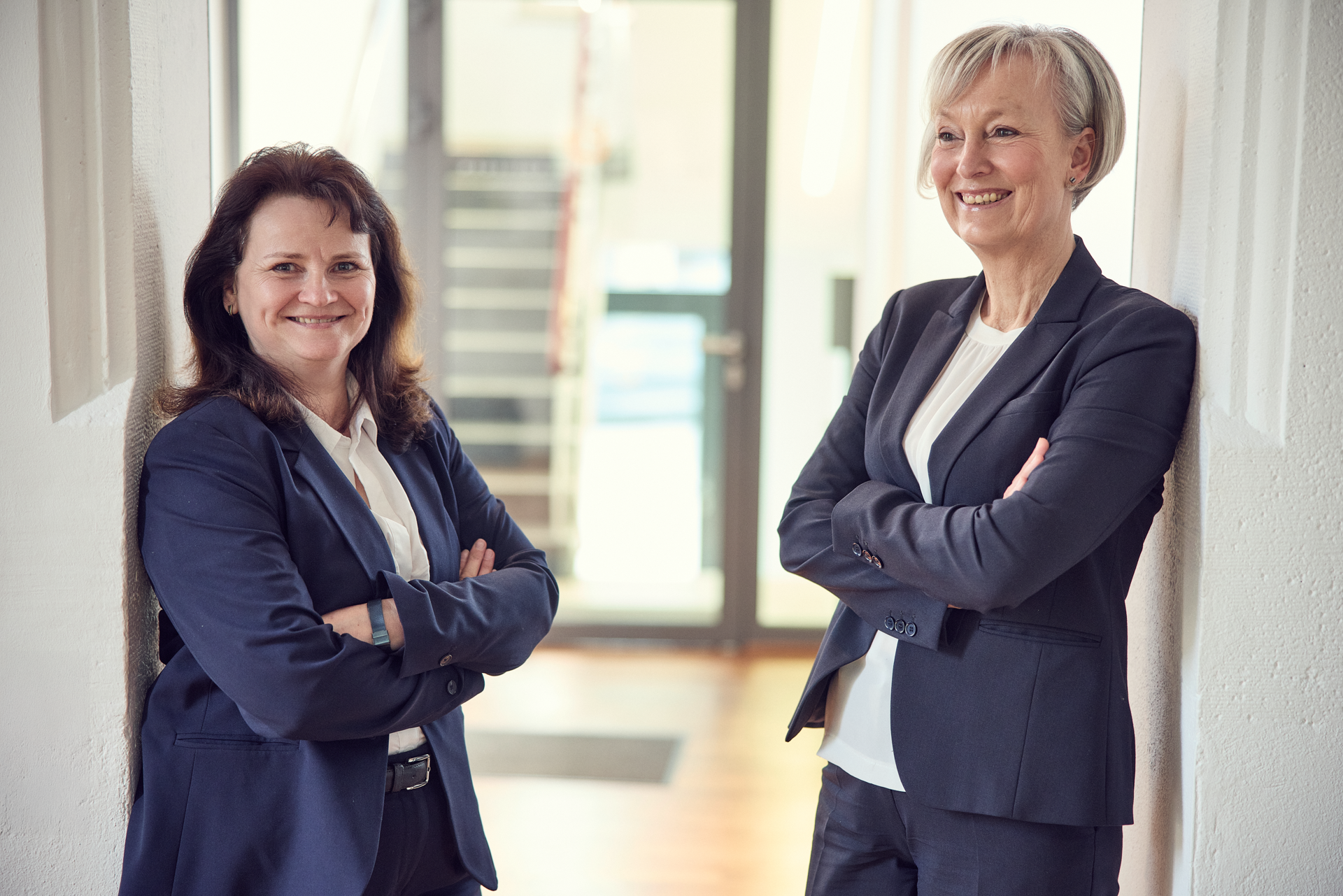 Zwei Frauen in formeller Kleidung stehen lächelnd nebeneinander in einem modernen Büroflur. Sie haben die Arme verschränkt und wirken freundlich und selbstbewusst. Im Hintergrund sind Treppen und ein Fenster zu sehen.