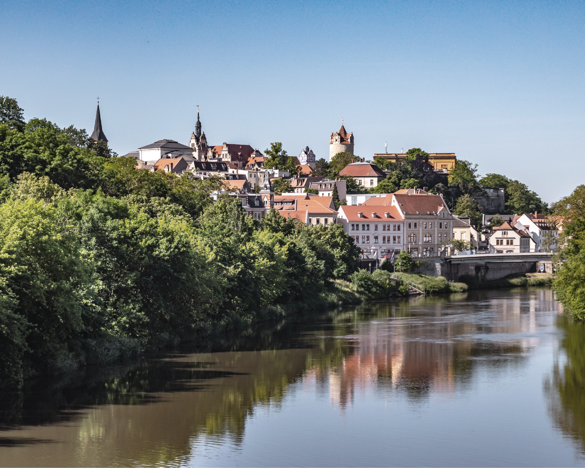 Eine malerische Aussicht auf eine Stadt, die am Ufer eines Flusses liegt, umgeben von grünen Bäumen und Hügeln, mit historischen Gebäuden und Kirchen im Hintergrund. Der Himmel ist klar und blau, während sich die Stadt im Wasser spiegelt.