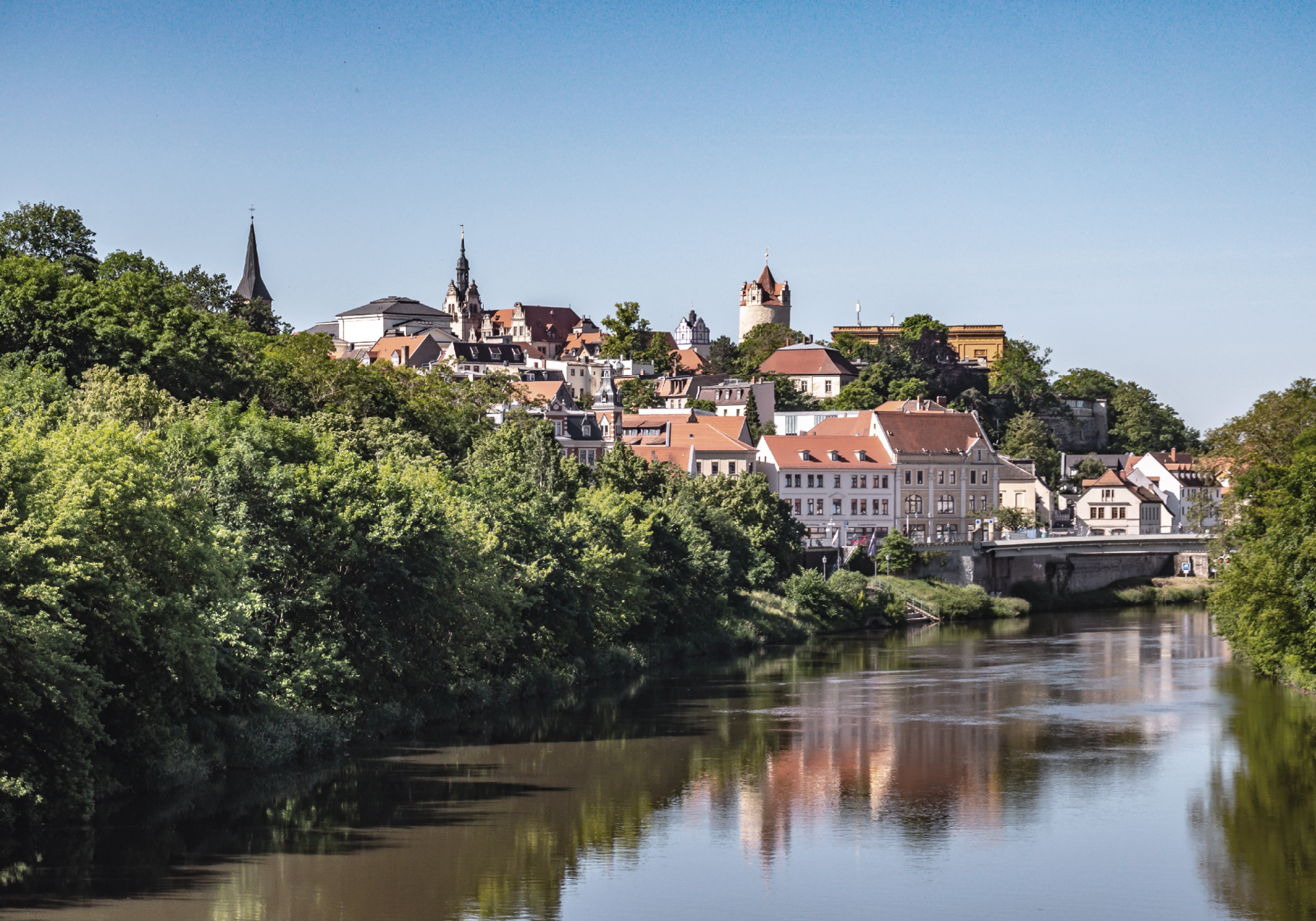 Eine malerische Stadtansicht mit bunt gedeckten Häusern und einem historischen Turm, umgeben von üppigem Grün und reflektiert im ruhigen Wasser eines Flusses.
