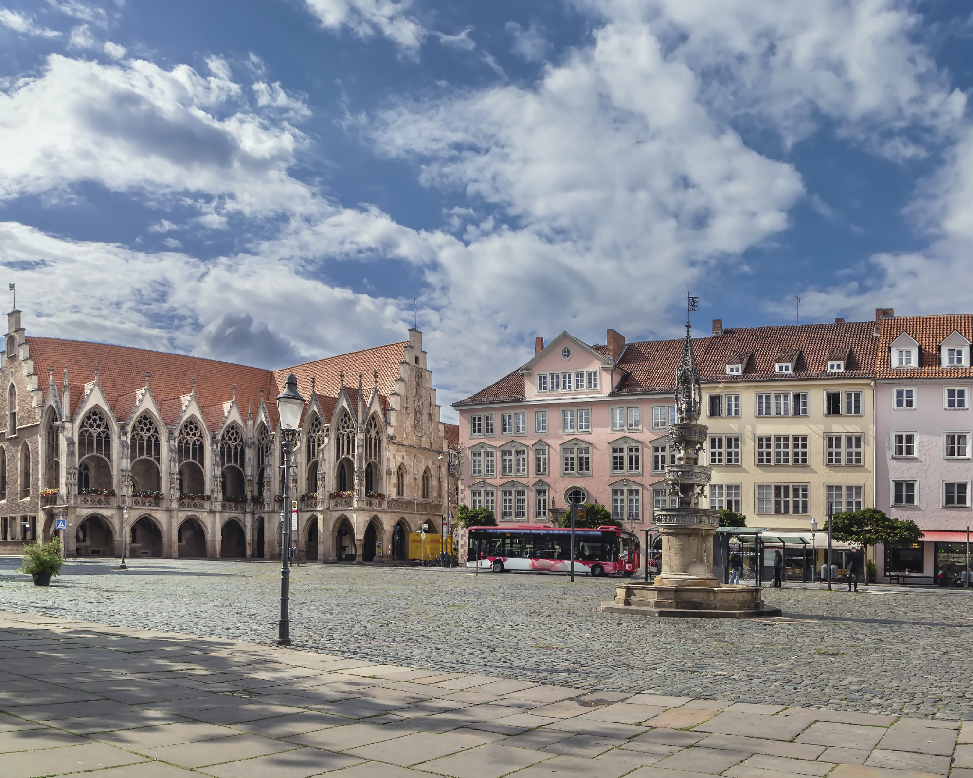 Der Marktplatz zeigt historische Gebäude mit einem gotischen Rathaus links, bunten Fassaden rechterhand und einer zentralen Brunnenanlage. Der Himmel ist mit einigen Wolken bedeckt, und ein Bus steht am Rand der Fläche.