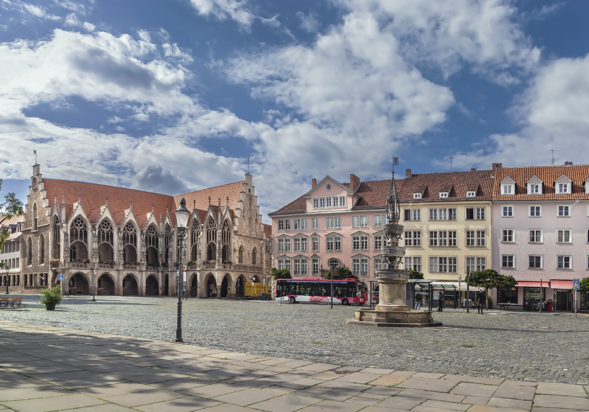 Blick auf einen historischen Platz mit gotischen Gebäuden, darunter ein Rathaus mit spitzbogen Fenstern, einer neugotischen Statue in der Mitte und pastelrosa Häusern im Hintergrund, unter einem klaren, bewölkten Himmel.
