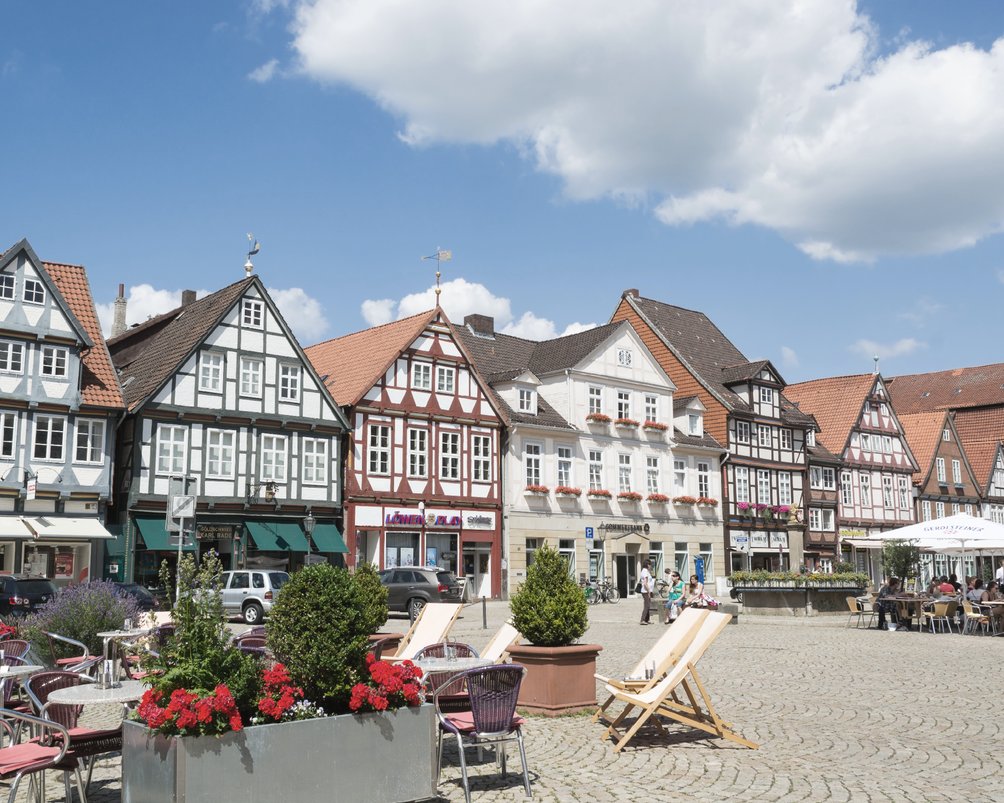 Ein malerischer Platz mit historischen Fachwerkhäusern, die in verschiedenen Farben gestaltet sind. Im Vordergrund befinden sich gemütliche Liegestühle und Blumenbeete mit bunten Blumen. Der Himmel ist blau mit einigen Wolken.