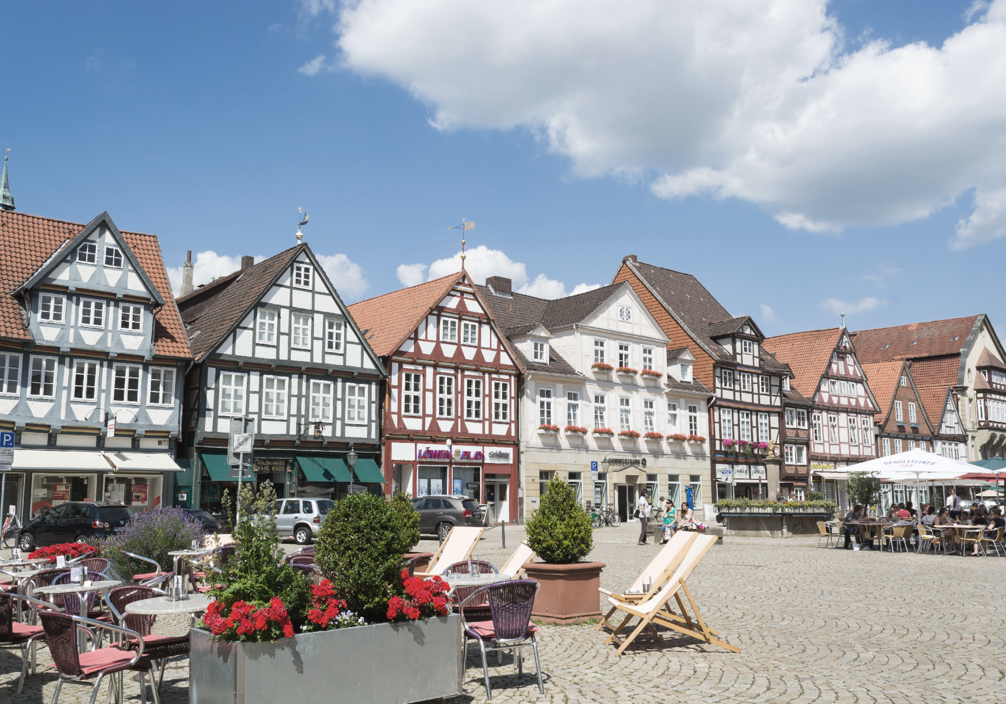 Der historische Marktplatz zeigt eine Reihe von bunten, zum Teil mit Fachwerk versehenen Gebäuden und gemütlichen Sitzgelegenheiten im Freien, umgeben von blühenden Pflanzen und einem klaren blauen Himmel.
