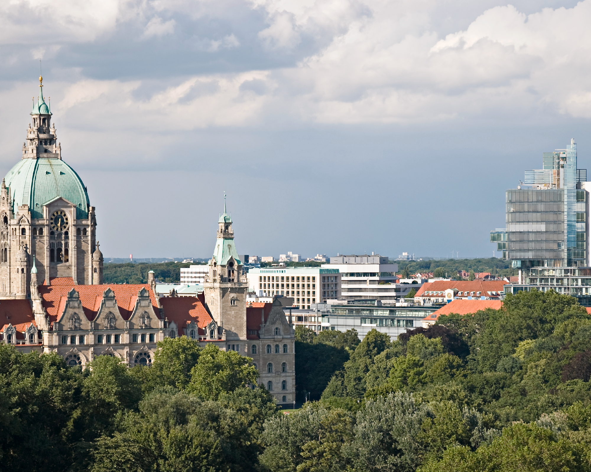 Die Stadtsilhouette von Hannover zeigt im Vordergrund das historische Rathaus mit einer markanten Kuppel, während im Hintergrund moderne Gebäude und eine bewaldete Landschaft zu sehen sind. Der Himmel ist bewölkt und gibt der Szene einen dramatischen Anstrich.