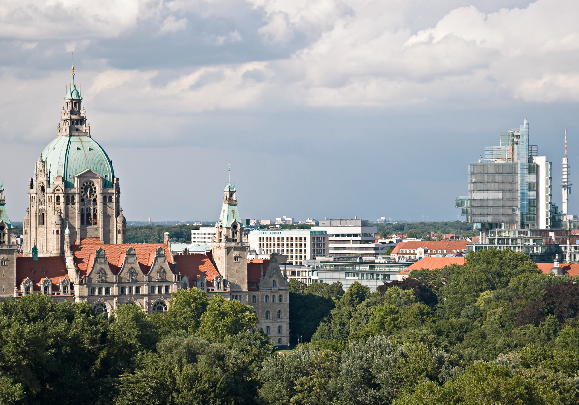 Die Skyline von Hannover mit dem historischen Rathaus im Vordergrund und modernen Bürogebäuden im Hintergrund, umgeben von Bäumen und einem bewölkten Himmel.