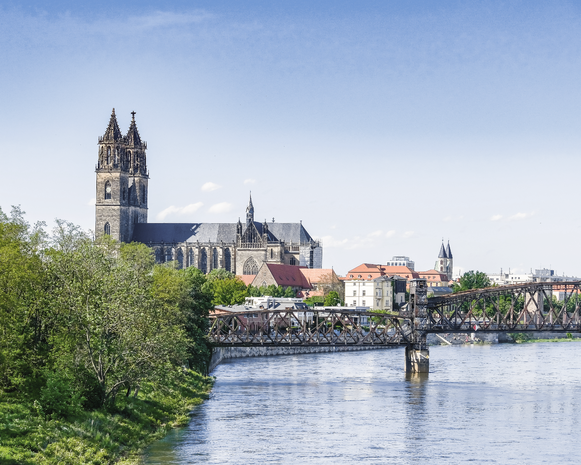 Die imposante Kirche mit einem hohen Turm überblickt eine Stadtlandschaft, während eine Eisenbahnbrücke über den Fluss führt und umliegendes Grün die Szenerie umrahmt.