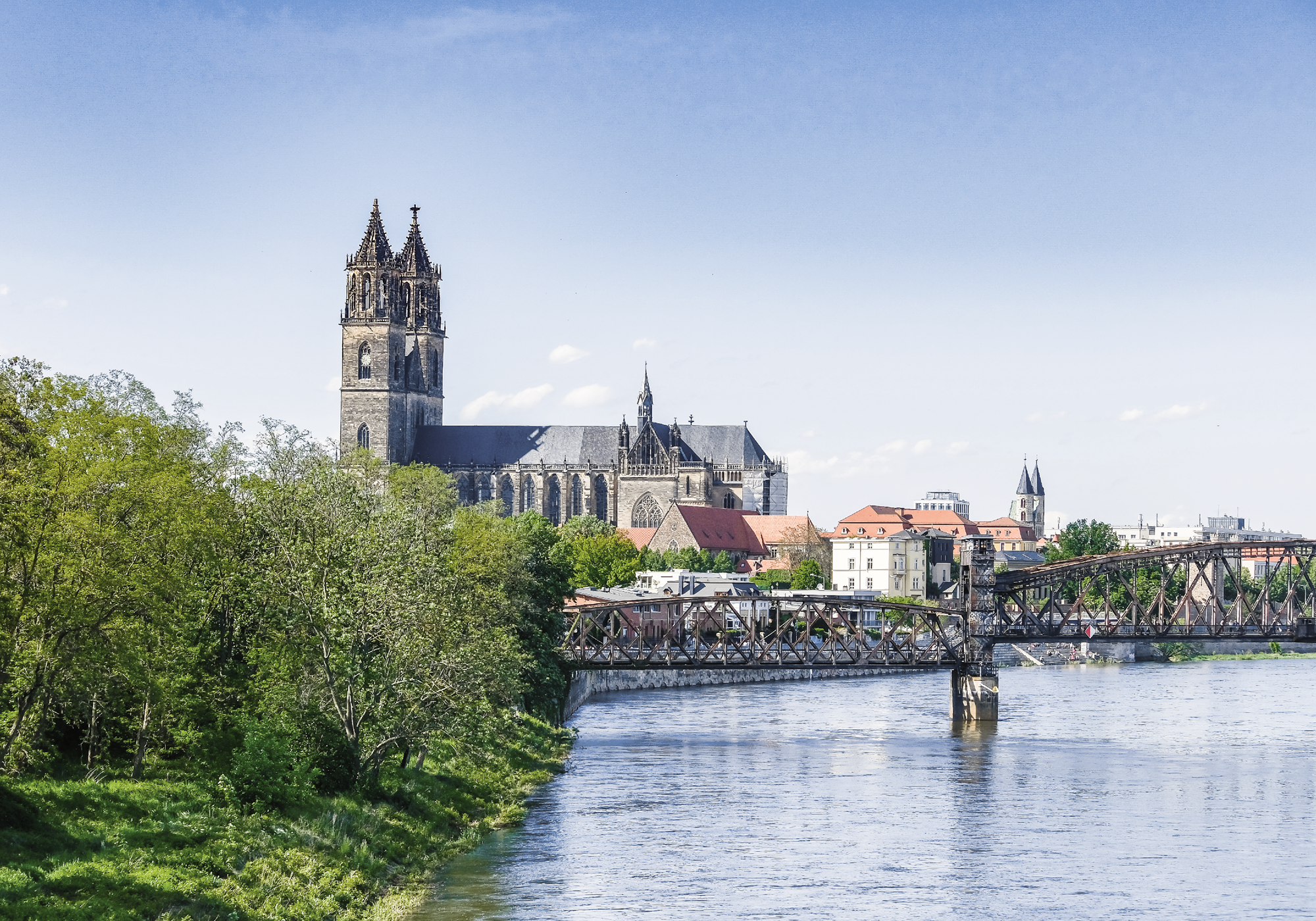Die Skyline einer Stadt mit einer markanten Kirche im Hintergrund, die von grünen Bäumen und einem Fluss umgeben ist, sowie einer Brücke, die über das Wasser führt.